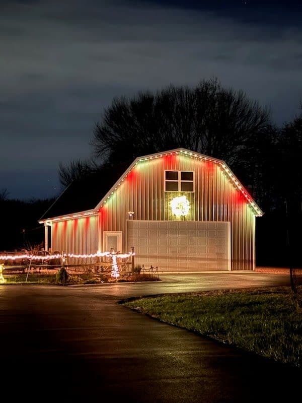 Christmas lights decorate a barn-style garage, featuring a wreath and red accents at night.