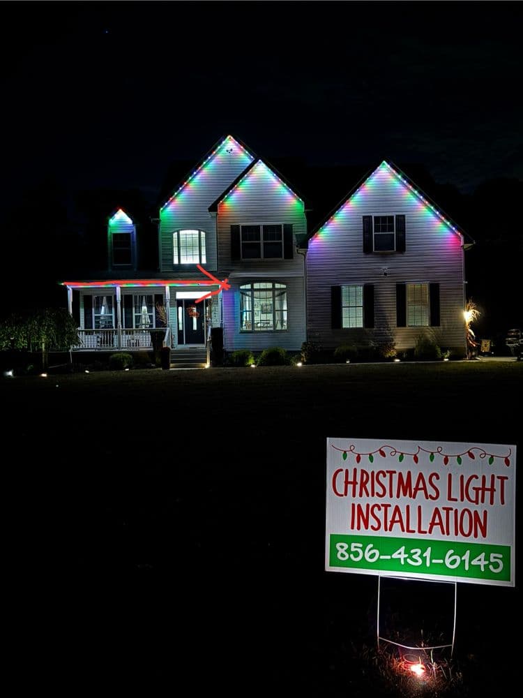 Christmas light installation on a home at night, featuring colorful lights and a promotional sign.