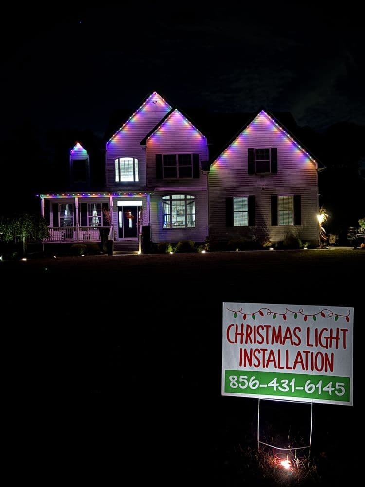 Christmas light installation on a decorated house at night with contact information sign.